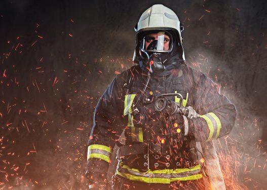 A professional firefighter dressed in uniform and an oxygen mask standing in fire sparks and smoke over a dark background. The image represents business owners firefighting with thier bookkeeping records.